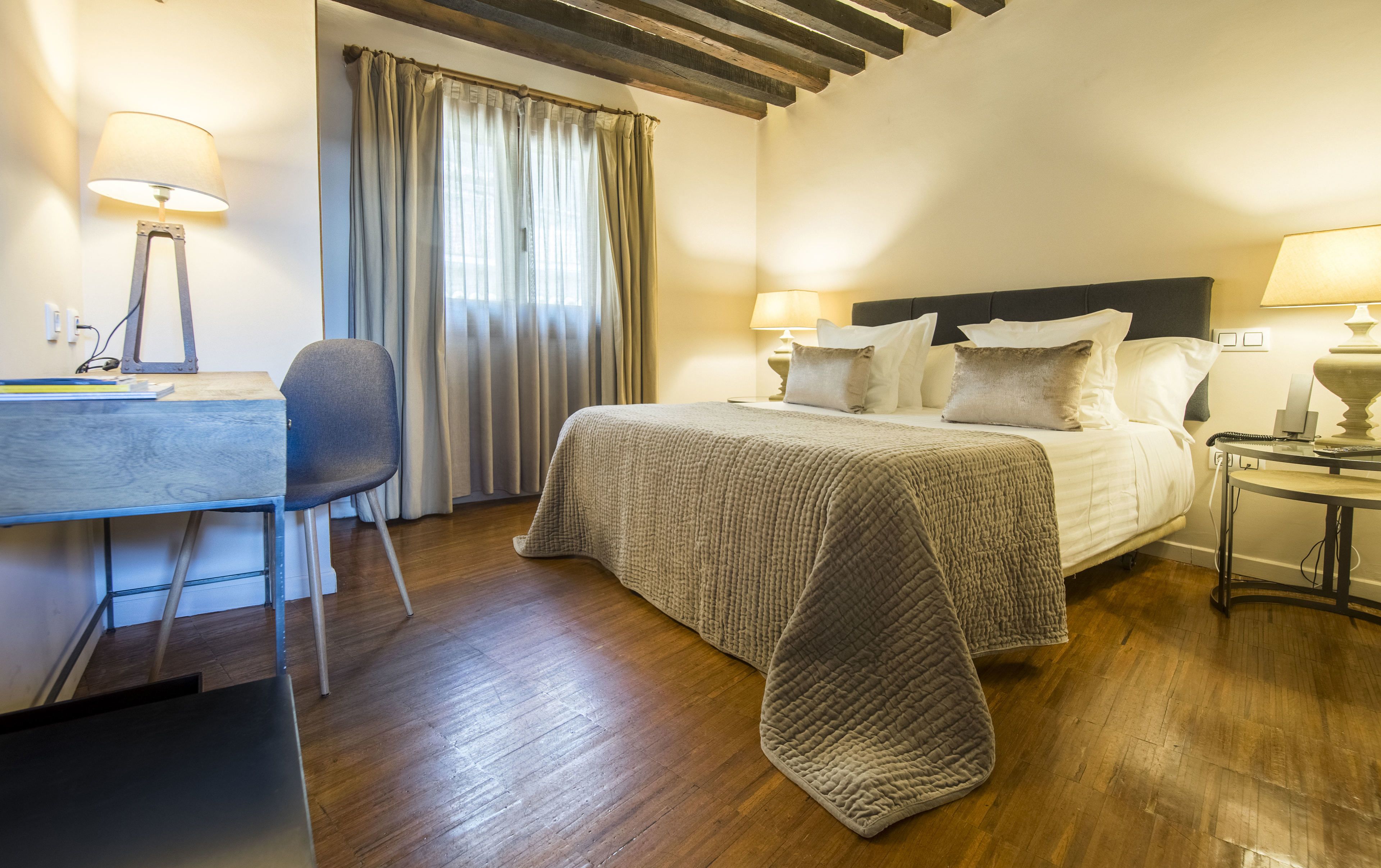 Una habitació d'hotel amb llit, làmpades de nit, escriptori i cadira, i finestra amb cortines.