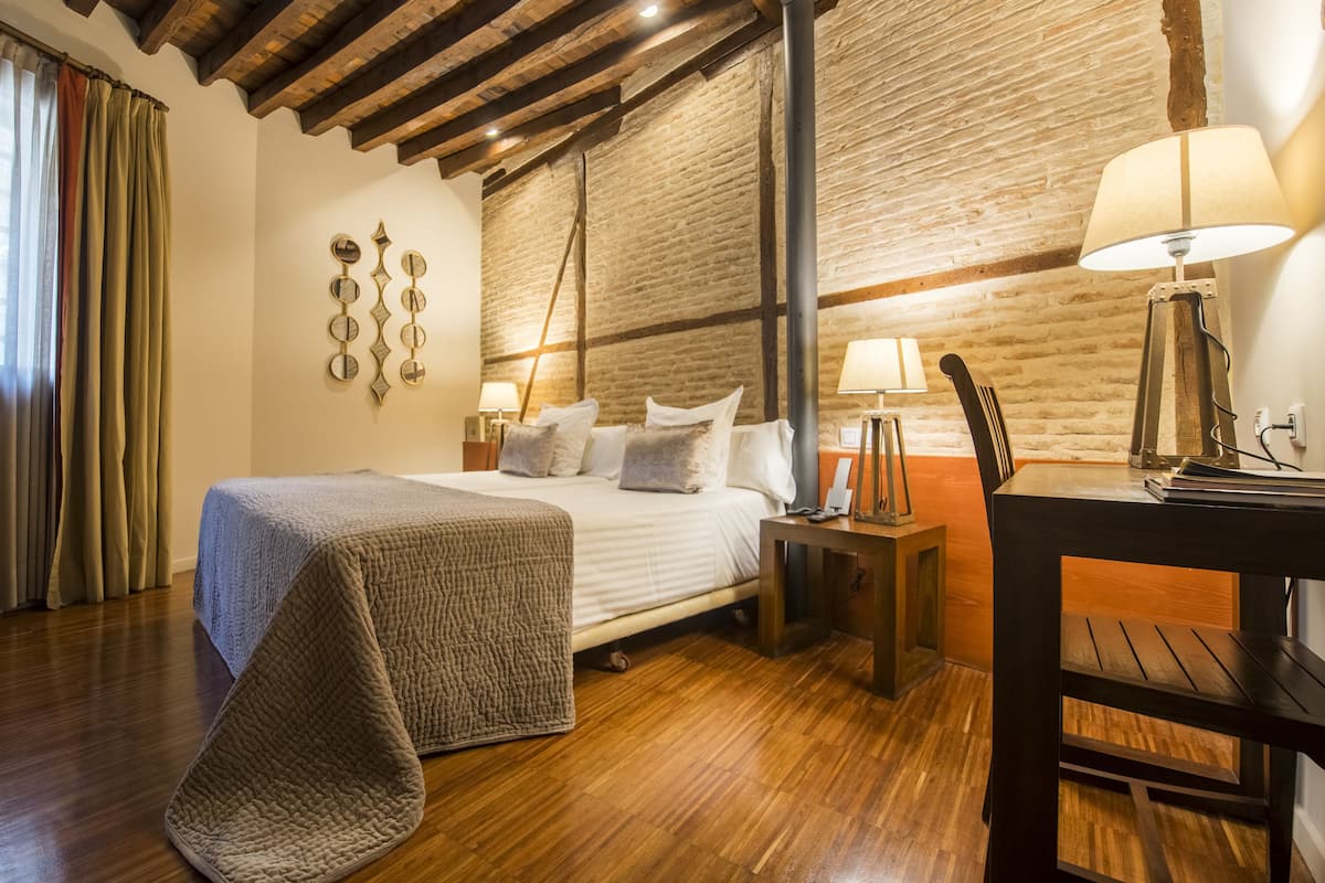 A bedroom with a wooden floor, a bed with a grey blanket, two bedside tables with lamps, a brick wall, and a wooden ceiling.
