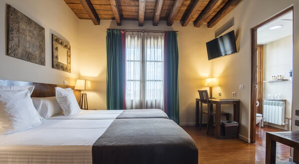 A hotel room with a double bed, a desk with a chair, a TV, and a window with curtains.