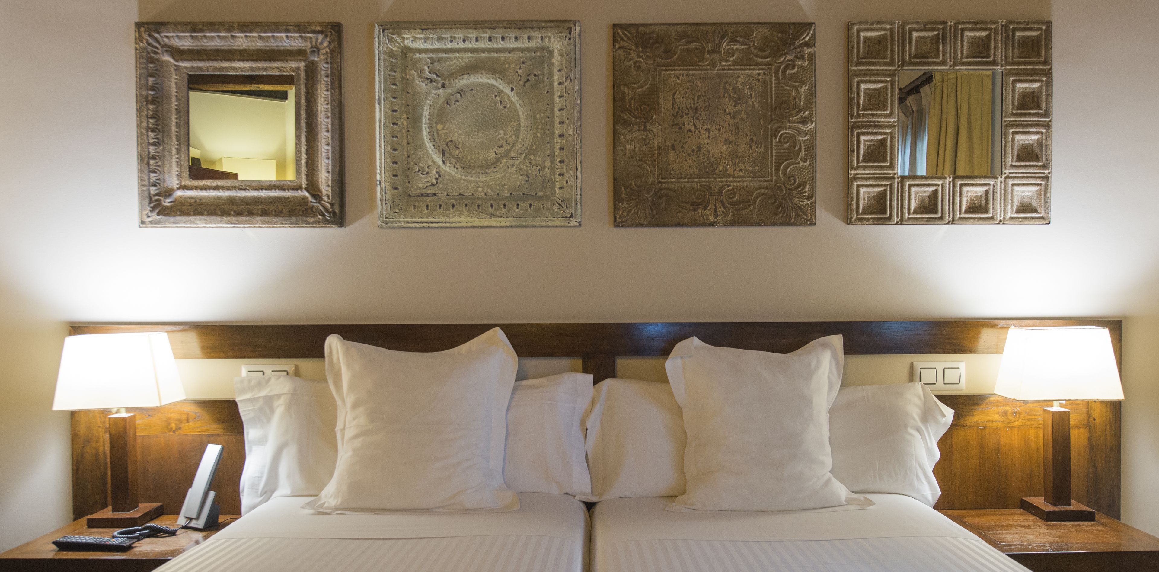 A hotel room with a double bed, two bedside tables with lamps, a telephone, and four framed artworks on the wall.