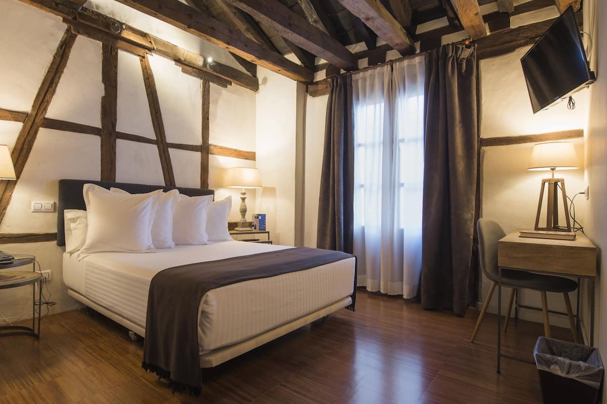 A hotel room with a wooden ceiling, a bed with white linens, two bedside lamps, a desk with a lamp, and a TV on the wall.