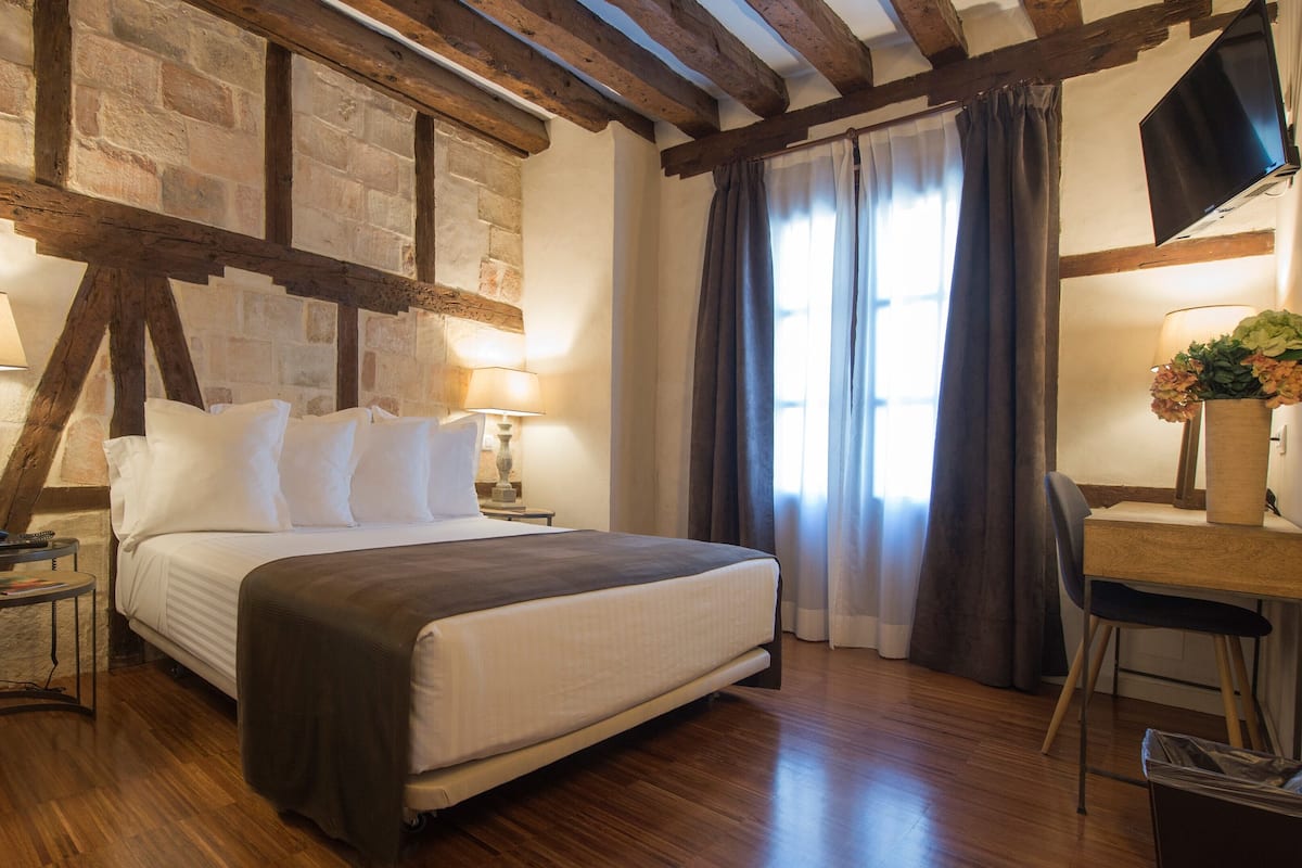 A hotel room with a wooden floor, a bed with white bedding, two bedside lamps, a desk with a chair, and a window with curtains.