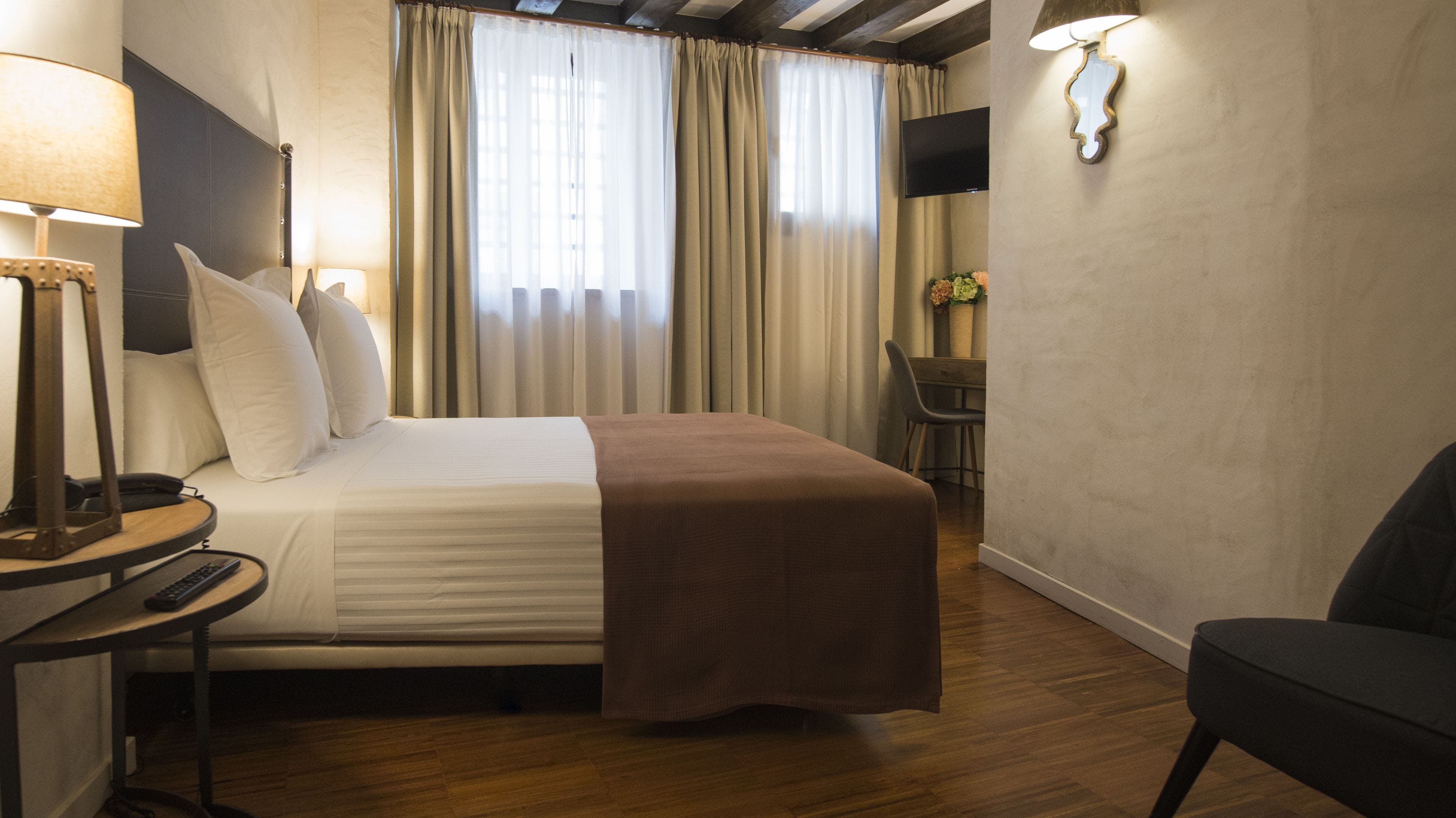 Habitación de hotel con cama, mesita de noche con lámpara, una silla, escritorio con silla y televisor montado en la pared.
