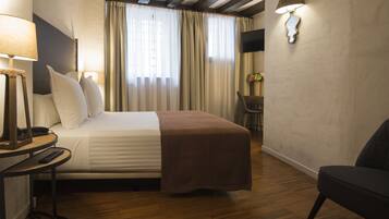 Habitación de hotel con cama, mesita de noche con lámpara, una silla, escritorio con silla y televisor montado en la pared.
