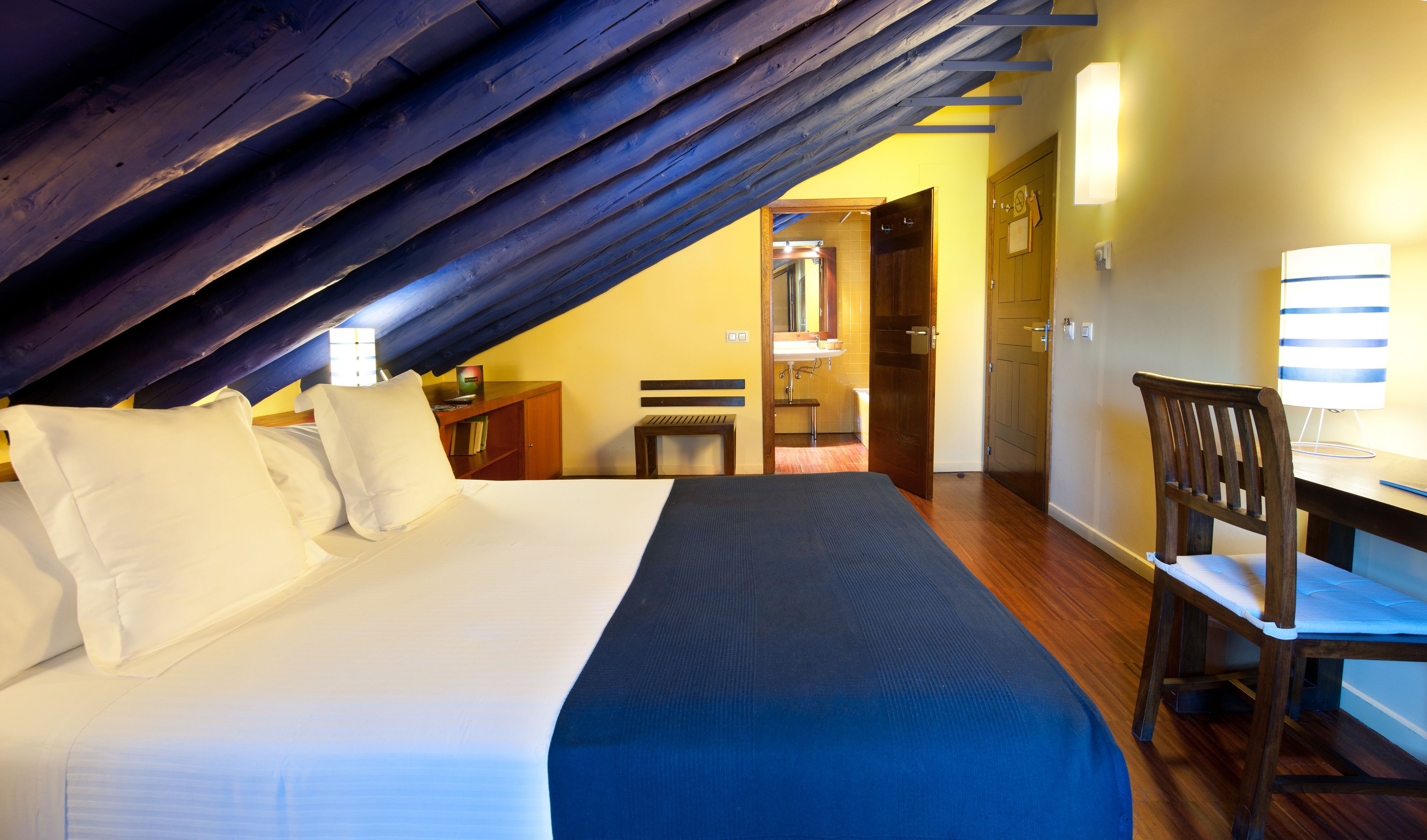 A bedroom with a large bed, wooden ceiling beams, a desk, and a chair.