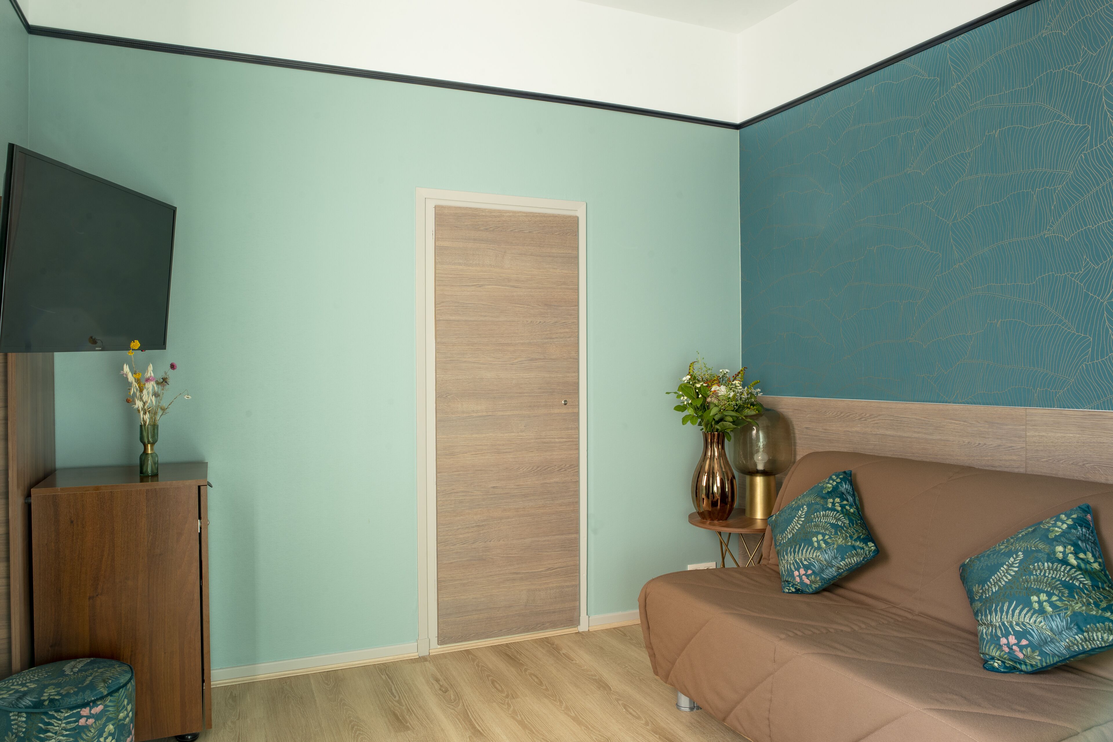 A room with a light blue wall, a wooden cabinet, a brown sofa with teal pillows, and a flat-screen TV mounted on the wall.