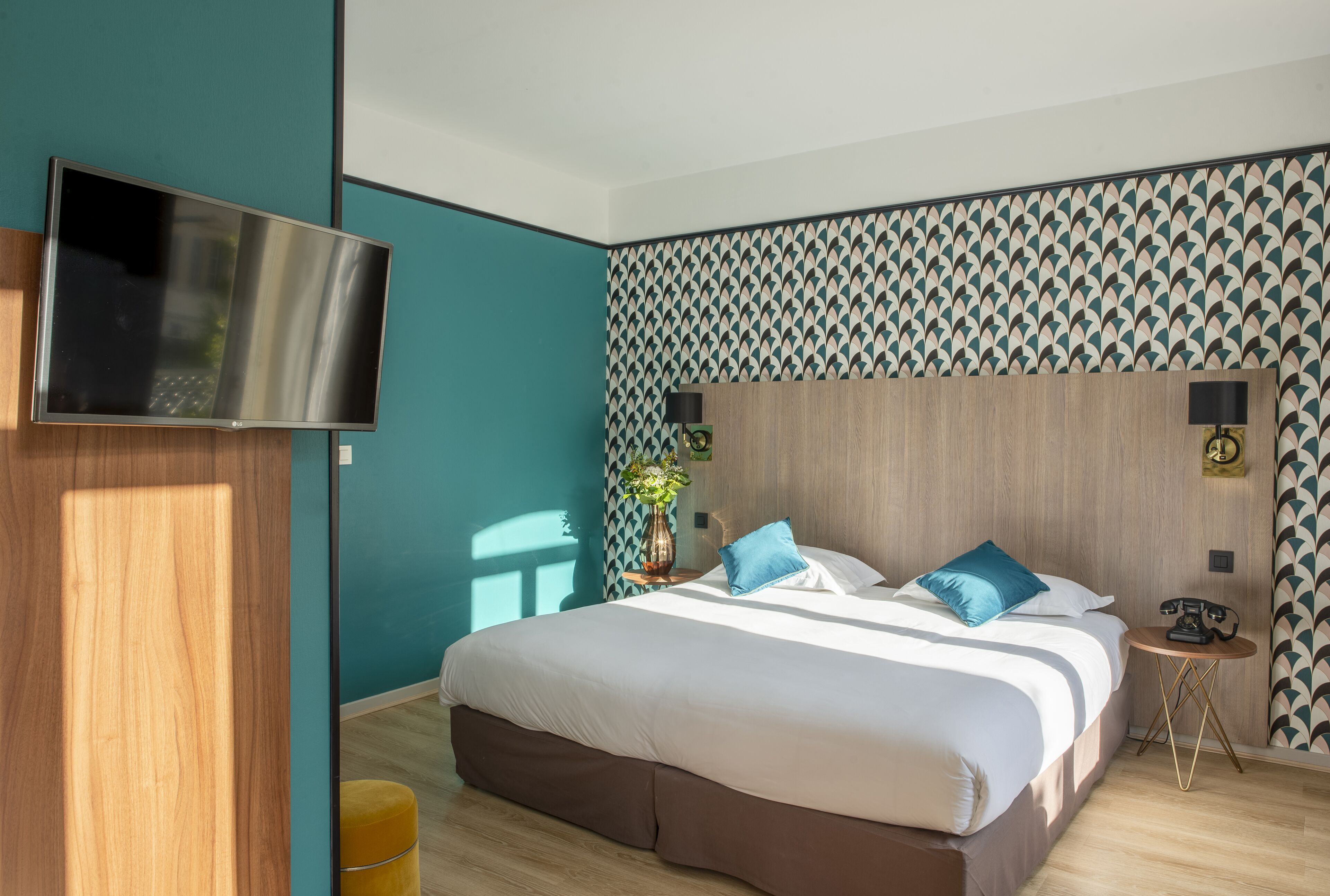 A modern hotel room with a large bed, a flat-screen TV mounted on the wall, and a patterned wallpaper.