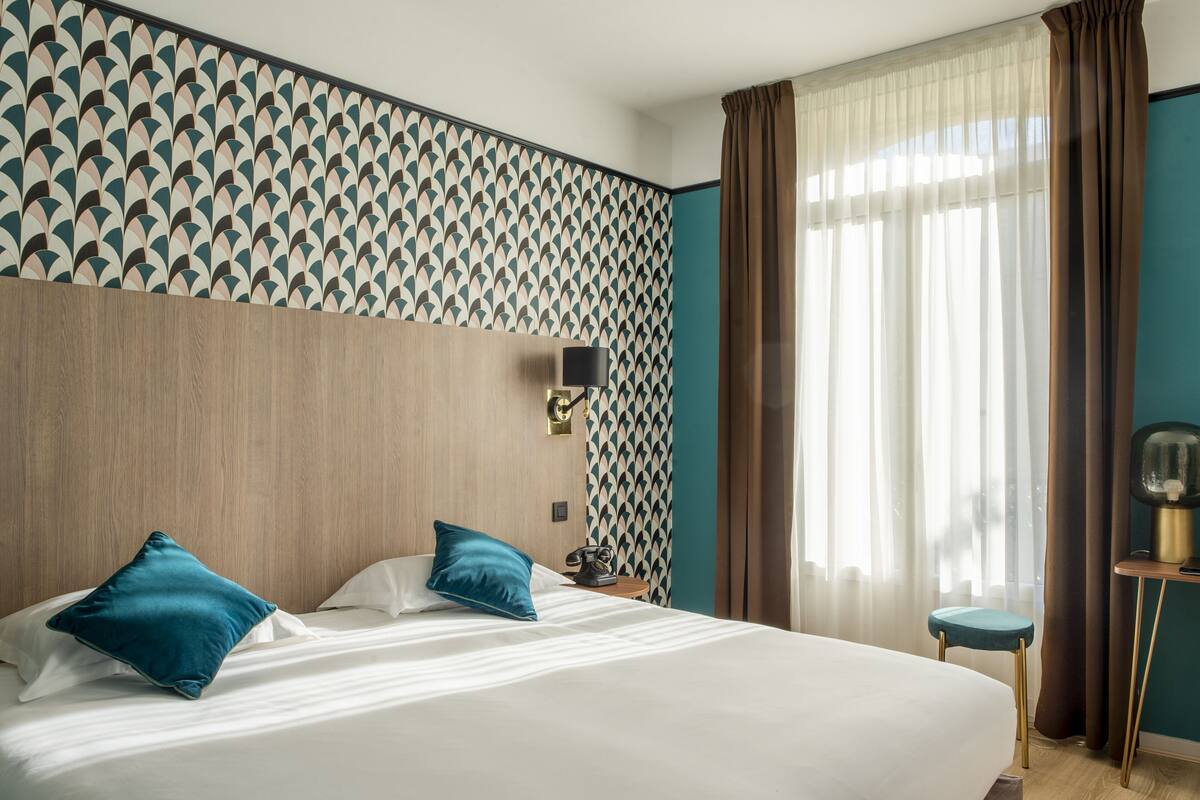 A hotel room with a large bed, a nightstand, a lamp, and a view of the outside through sheer curtains.