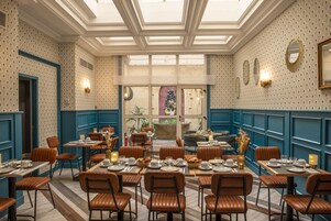 A restaurant with a high ceiling, patterned wallpaper, and blue paneling. Tables are set with plates, glasses, and candles.