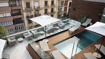 A rooftop pool area with lounge chairs and umbrellas, surrounded by a wooden deck and glass railings.