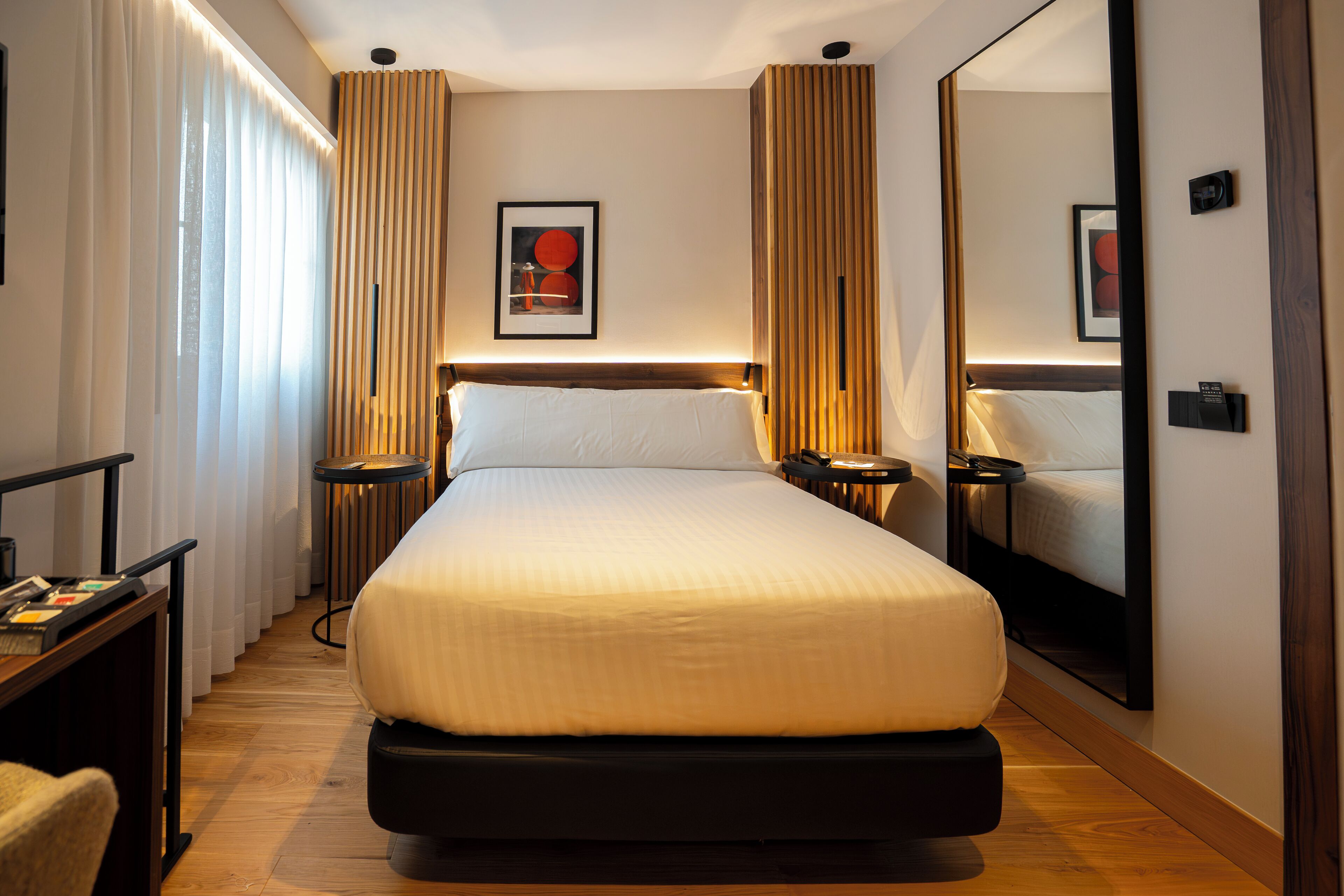 Habitación de hotel con una cama grande, un escritorio y una silla. Hay un espejo en la pared y un cuadro enmarcado sobre la cama.