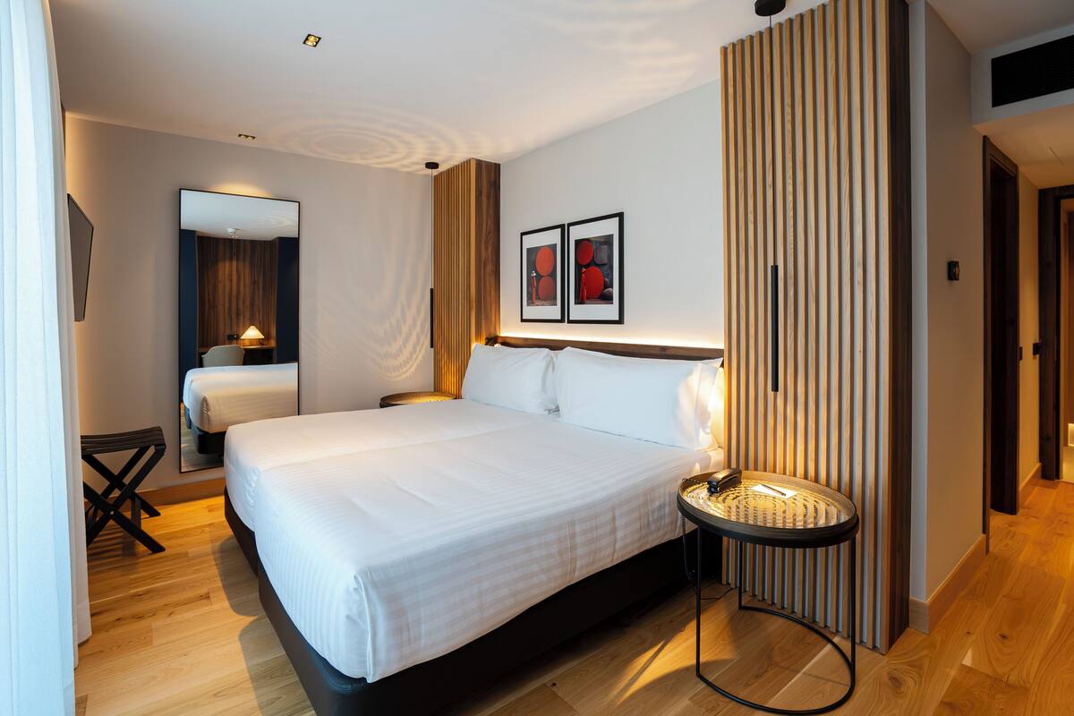 Moderni hotellihuone, jossa on suuri sänky, pieni pyöreä pöytä, tuoli, peili ja kaksi kehystettyä kuvaa seinällä.