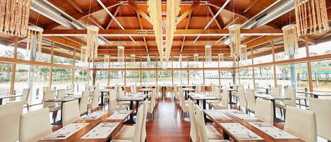 A spacious indoor dining area with wooden tables and chairs, large windows, and hanging light fixtures.