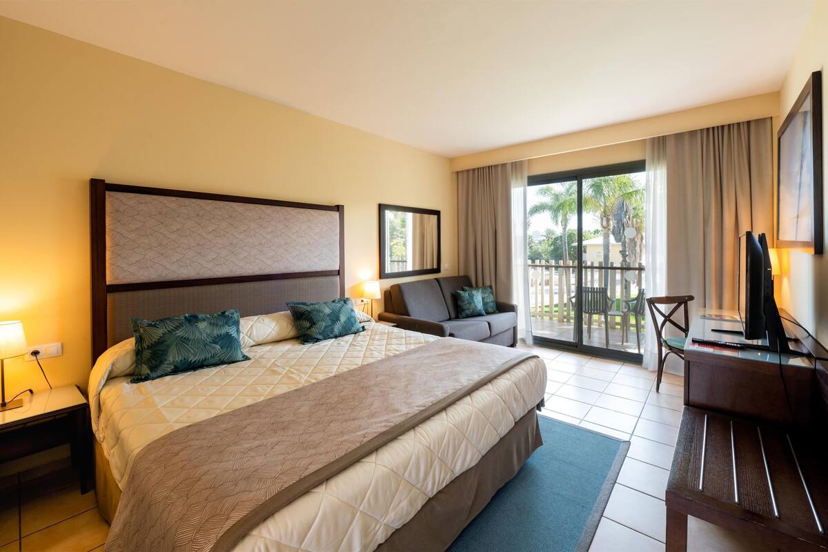 Habitación de hotel con una cama grande, un escritorio con computadora, una silla y un balcón con vista a palmeras.