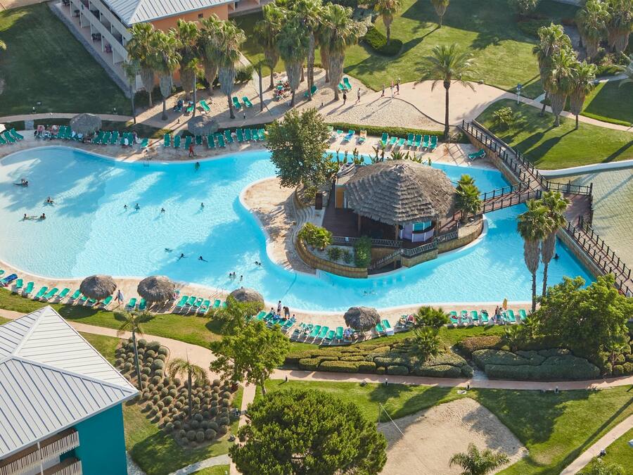 Eine Luftaufnahme eines Resorts mit einem zentralen Schwimmbecken, mehreren Liegebereichen und einem strohgedeckten Gebäude.