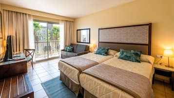Habitación de hotel con dos camas, un escritorio, una silla y un balcón con vistas a palmeras.