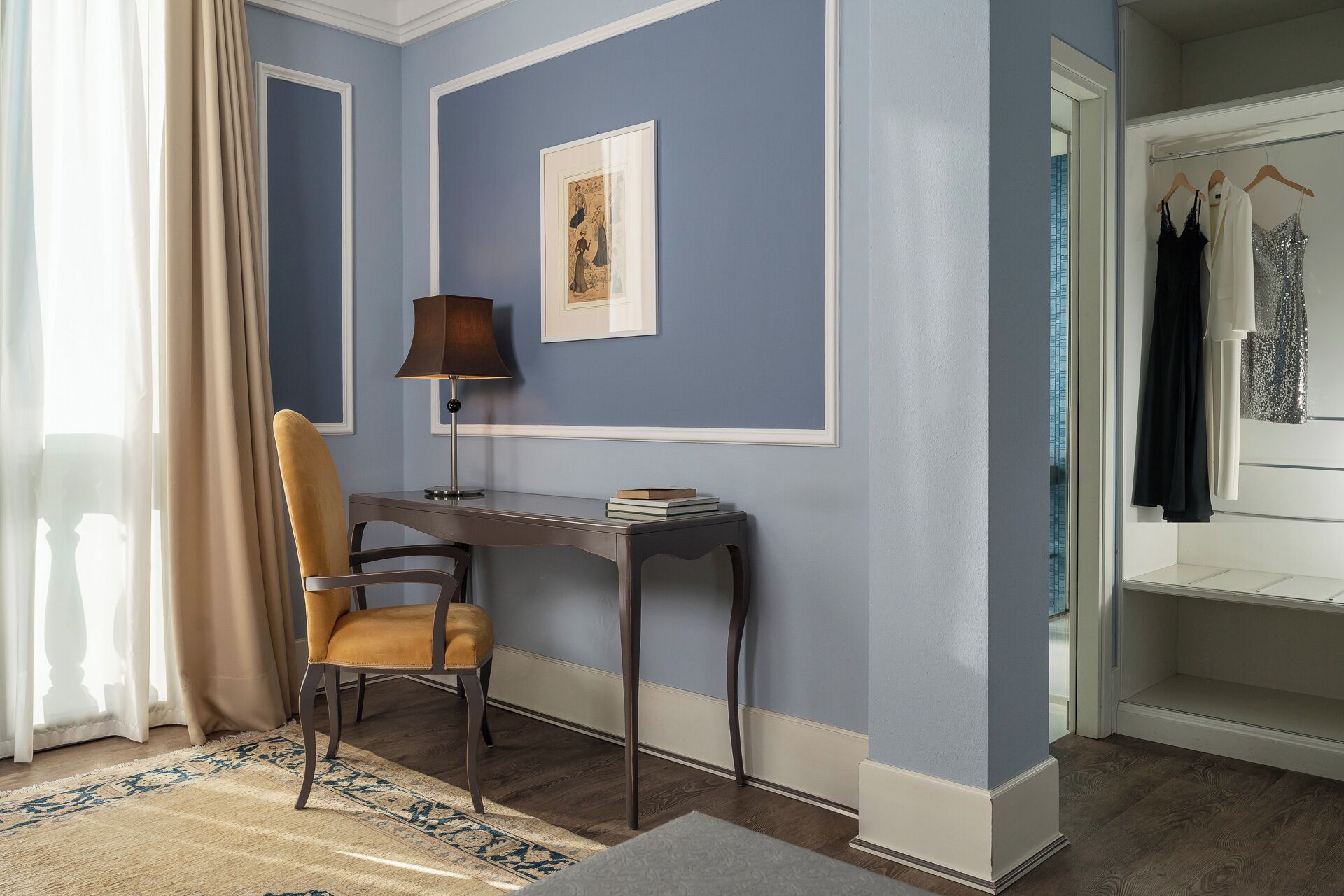 A room with a desk, chair, lamp, framed picture, and a clothing rack.