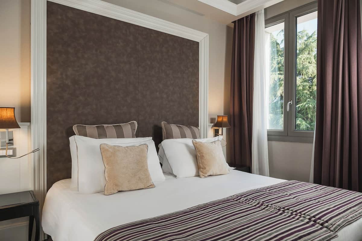 A hotel room with a large bed, two bedside lamps, a window with curtains, and a view of trees.