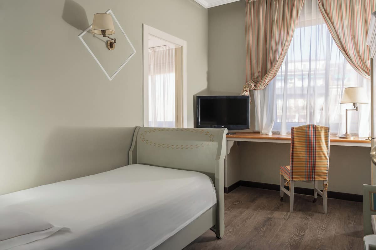 A hotel room with a single bed, a desk with a television, a chair, and a window with curtains.