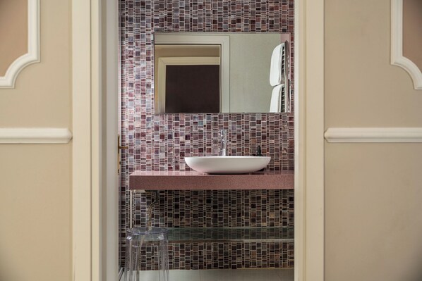 Un baño con un lavamanos blanco tipo recipiente, un taburete de cristal y un salpicadero de azulejos.