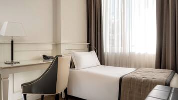A hotel room with a bed, a desk with a lamp, a chair, and a window with curtains.