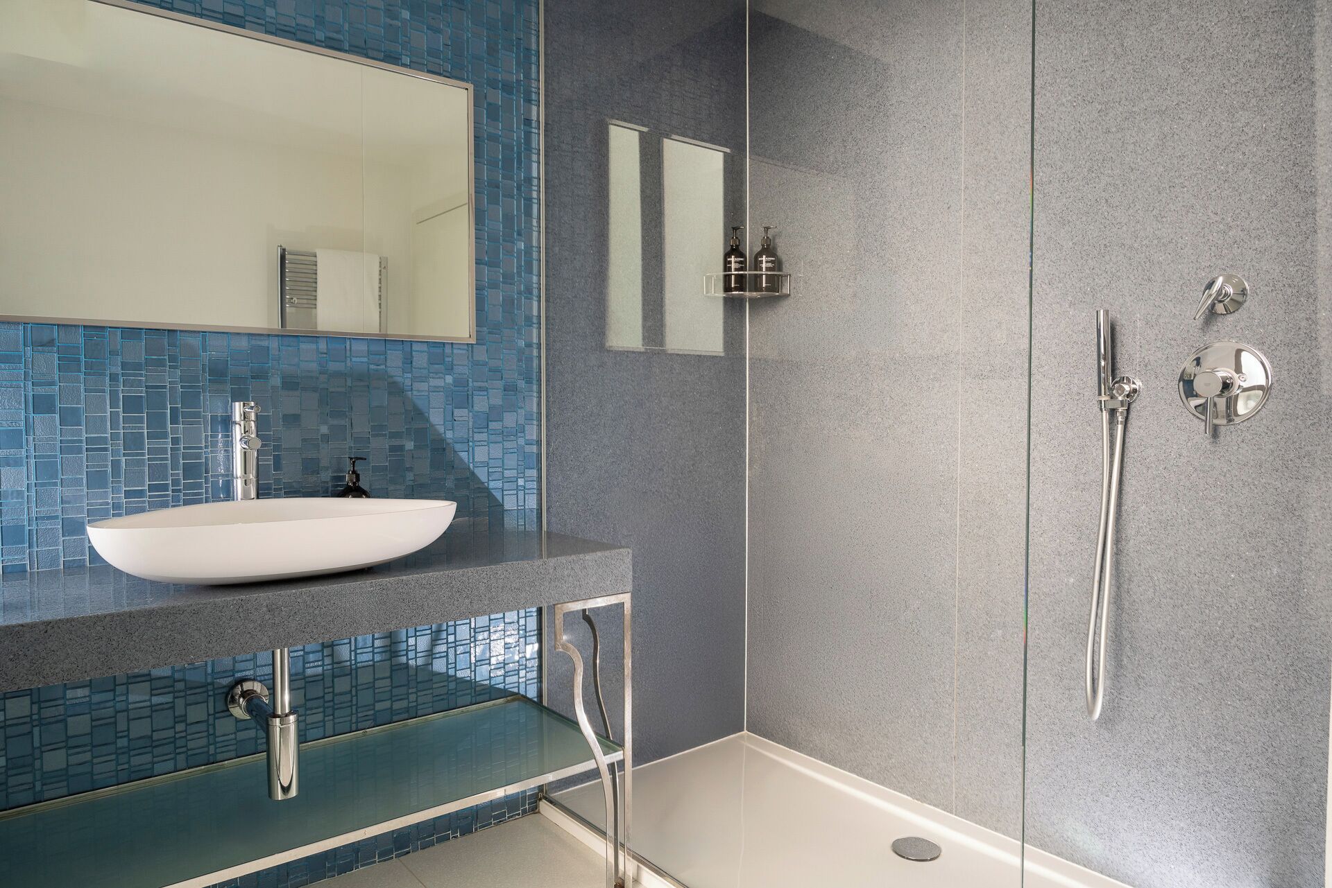 Un baño moderno con un lavamanos blanco, mampara de ducha de vidrio y salpicadero de mosaico azul.