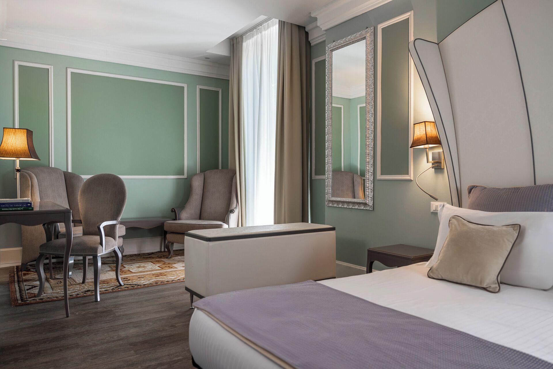 Habitación de hotel con cama, escritorio, dos sillas, un espejo grande y una mesita de noche.