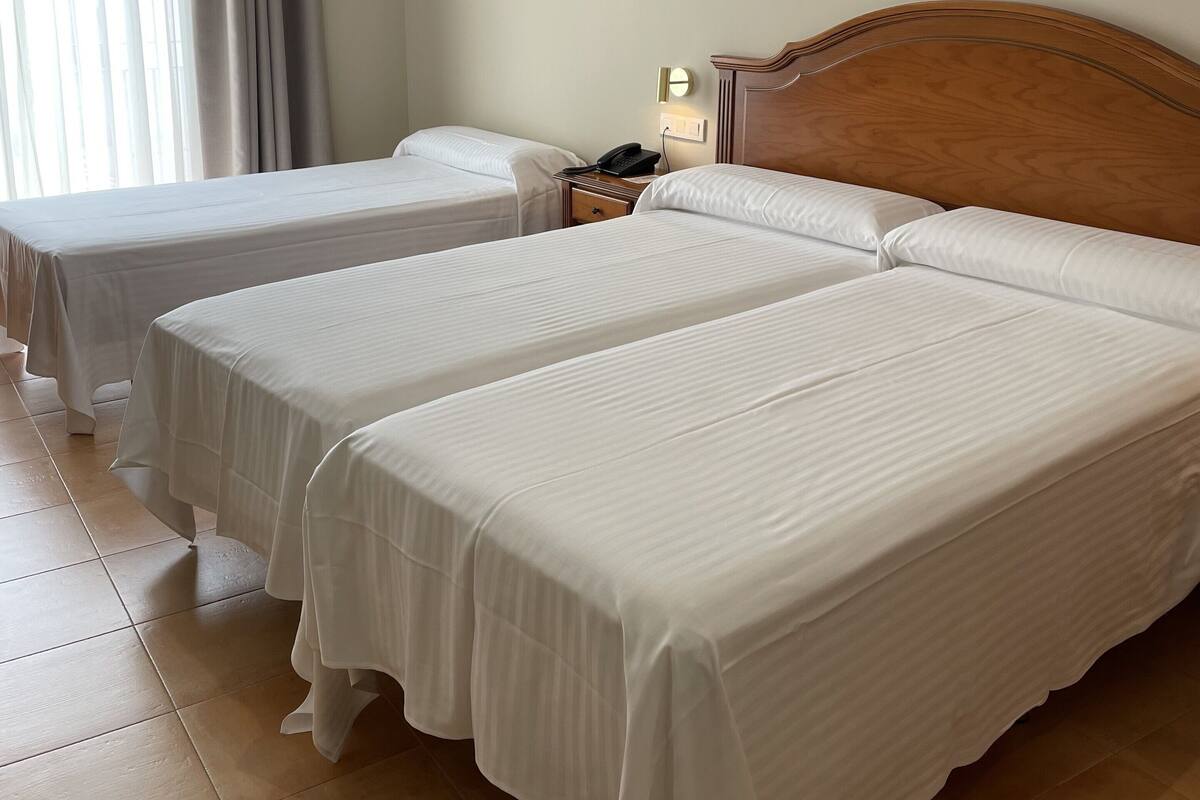 A hotel room with two beds, a wooden headboard, a bedside table with a lamp, and a window with curtains.
