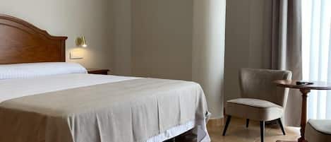 A hotel room with a large bed, a chair, a small table, and a window with curtains.