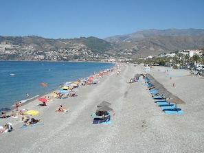 A beach with rows of umbrellas and sun loungers, a clear blue sea, and a hilly town in the background.