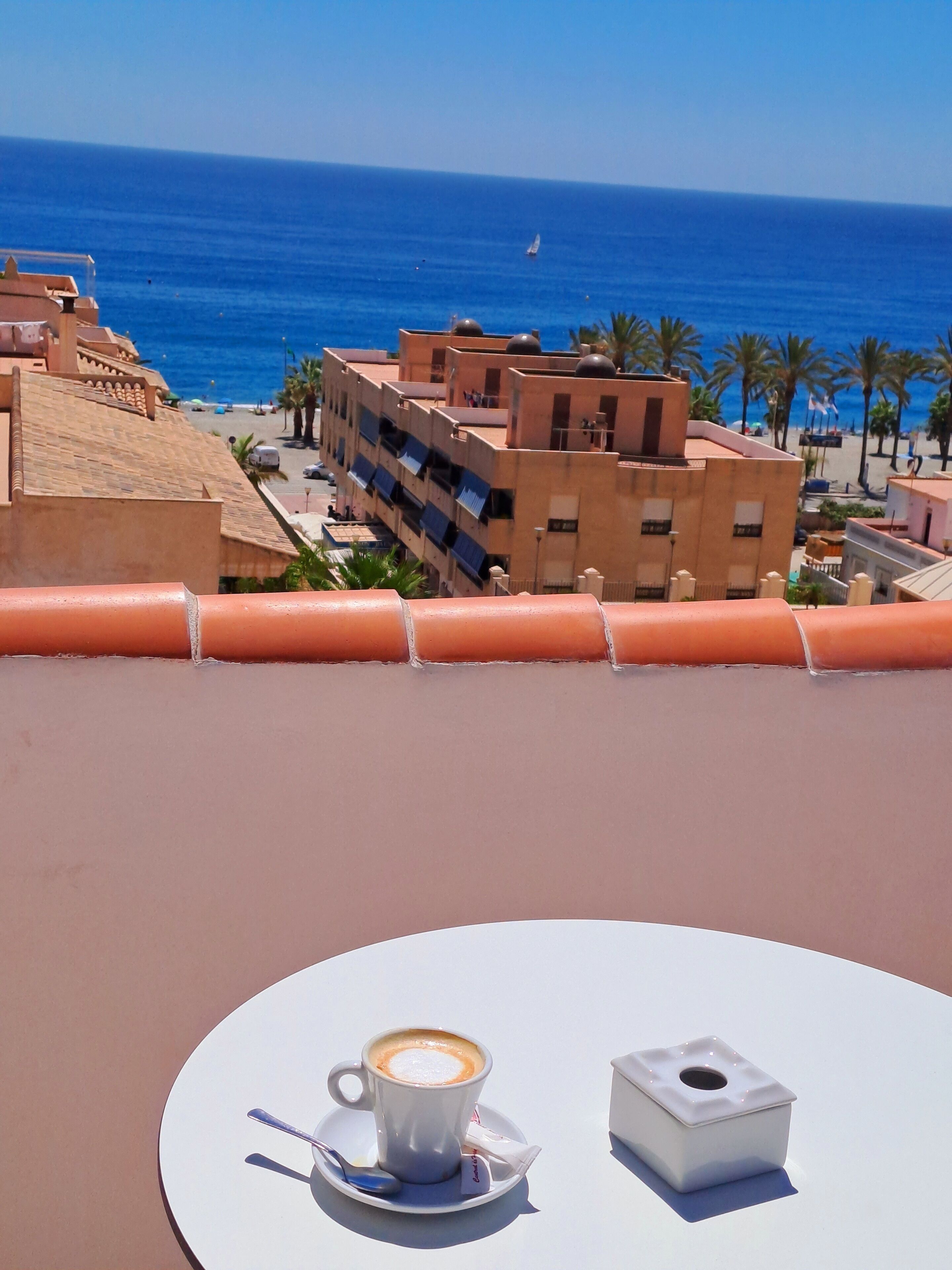 A table with a cup of coffee and a box, overlooking a coastal view with buildings and palm trees.
