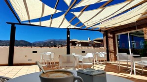 A table with a cup labeled "SOLOCREMA" and a plate, under a white and blue canopy, with a view of mountains and a building in the background. - Hotel Almijara – Mares (Almunecar)