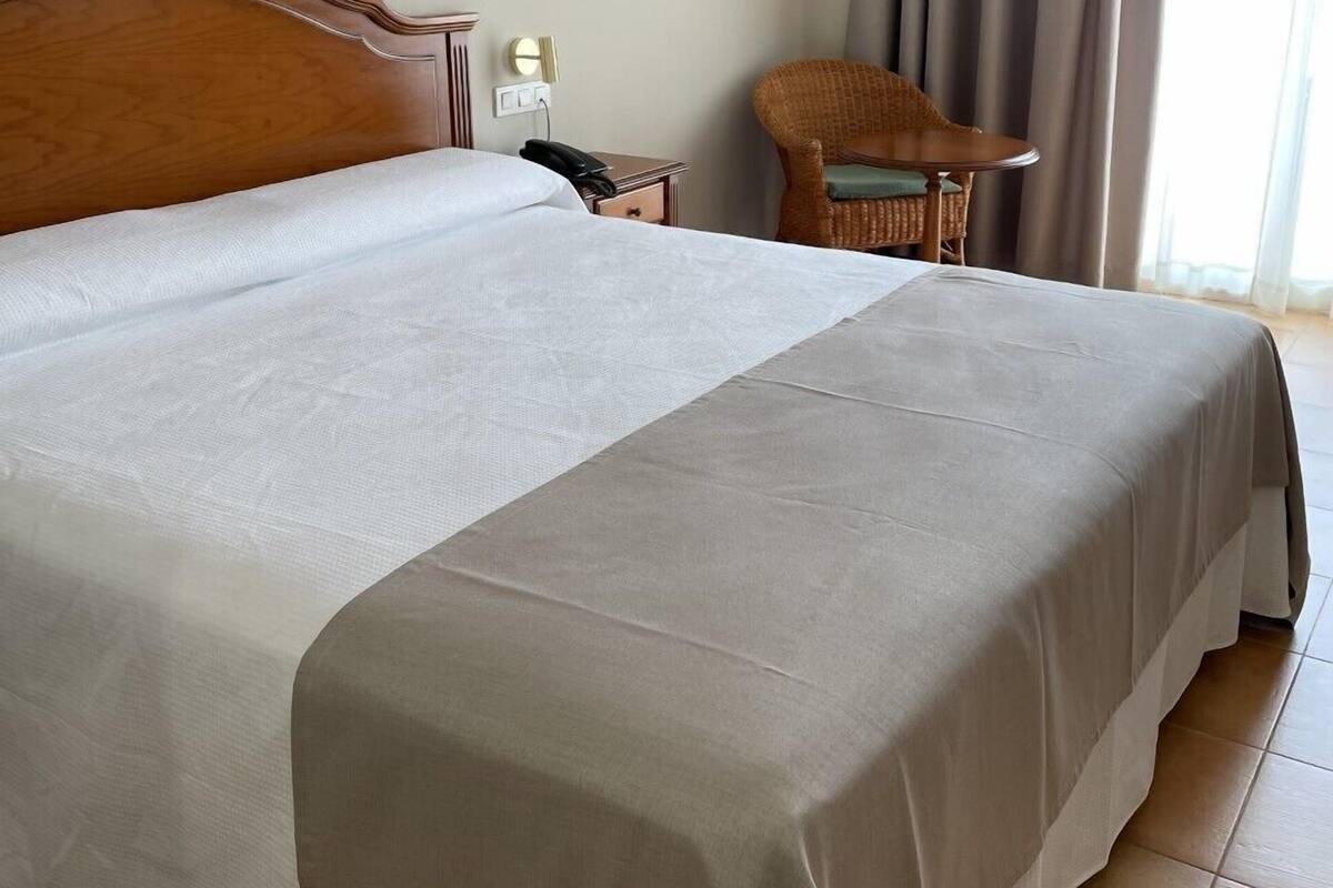 A hotel room with a large bed, a wooden headboard, a bedside table with a lamp, a chair, and a window with curtains.