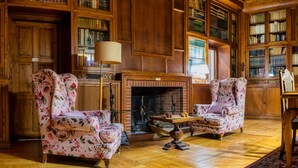 A library with wooden bookshelves, two floral armchairs, a fireplace, and a wooden floor. - Els Jardins De La Martana (Besalu)