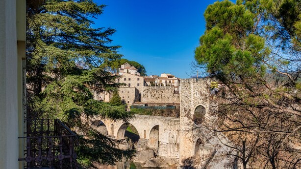 A stone bridge with arches, a river below, and a building with multiple levels in the background. - Els Jardins De La Martana (Besalu)