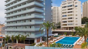 Un hotel de varios pisos con piscina, palmeras y otros edificios al fondo.