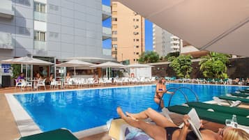 Uma área de piscina com espreguiçadeiras e guarda-sóis, rodeada por edifícios modernos.