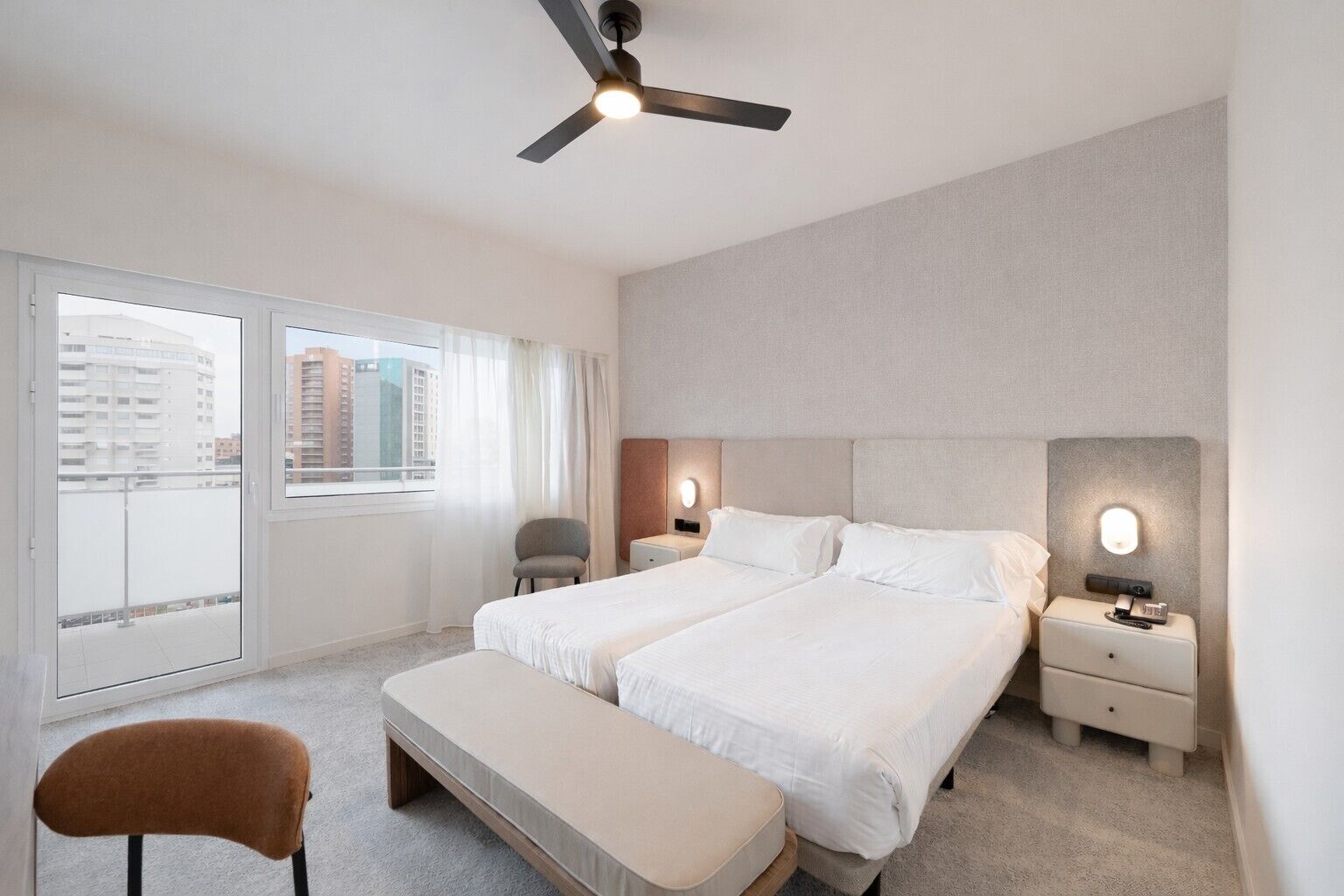 Een hotelkamer met twee bedden, een plafondventilator, een bank, een stoel en uitzicht op gebouwen door het raam.