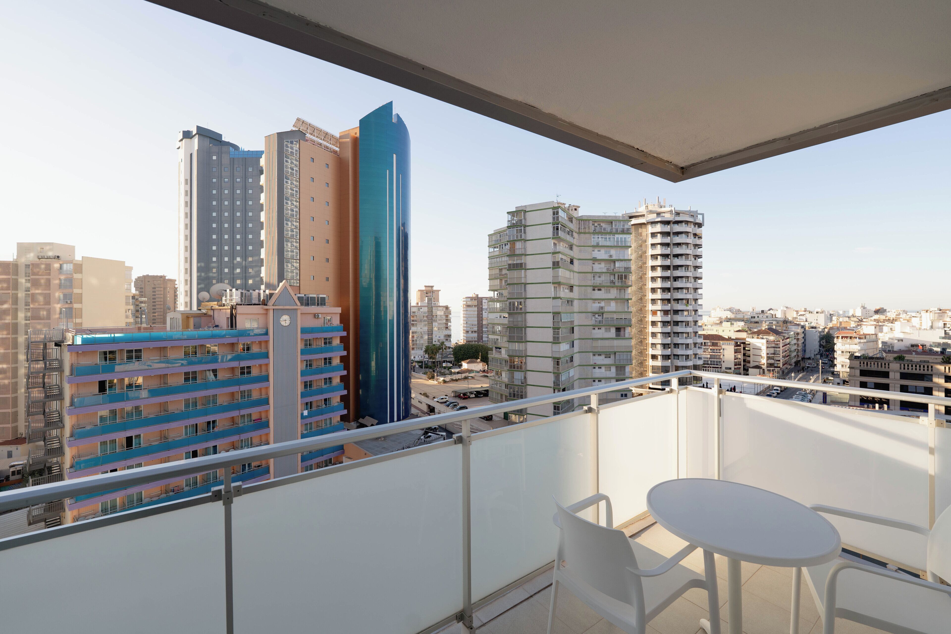 Un balcón con una mesa y sillas blancas, con vista a un paisaje urbano con varios edificios.