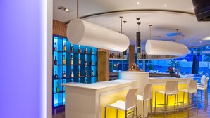 Un área de bar moderna con taburetes blancos, un mueble iluminado en azul con botellas y un diseño de techo curvo.