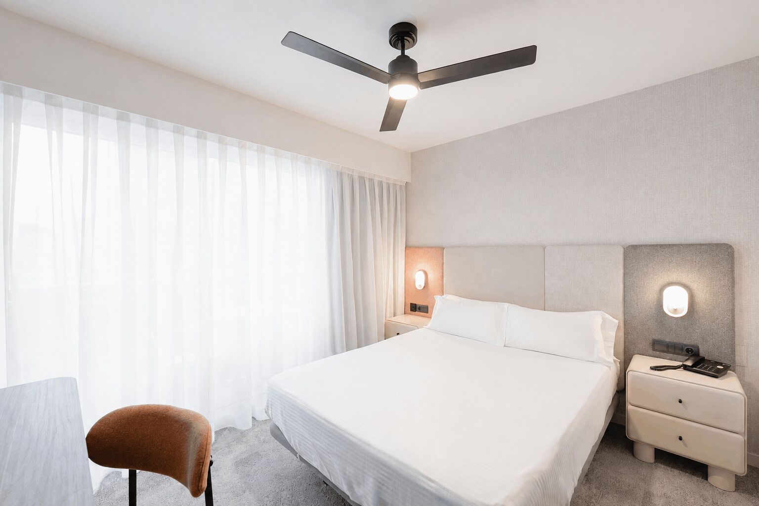A hotel room with a bed, a desk, a chair, a ceiling fan, and two bedside tables with lamps.