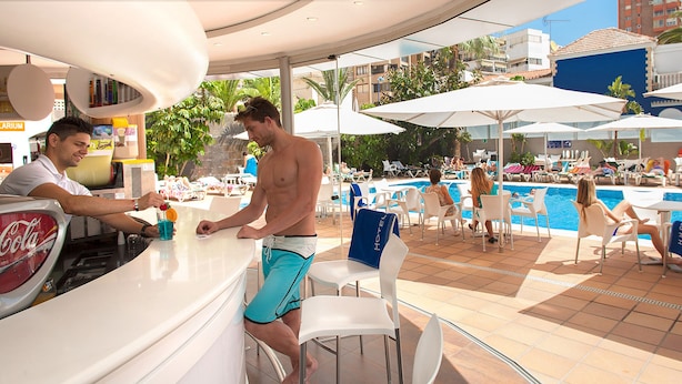 Zona de bar junto a la piscina con refrigerador de Coca-Cola, taburetes blancos y piscina con sillones y sombrillas.
