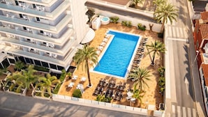 Una piscina en la azotea con tumbonas y palmeras, rodeada de edificios altos.