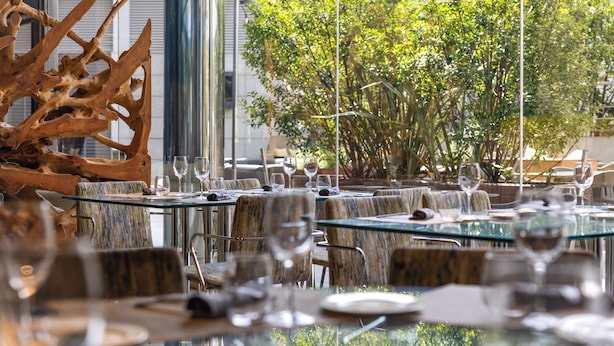 Un restaurant avec des tables et des chaises en verre, une grande sculpture en bois et une vue sur des arbres et un bâtiment.