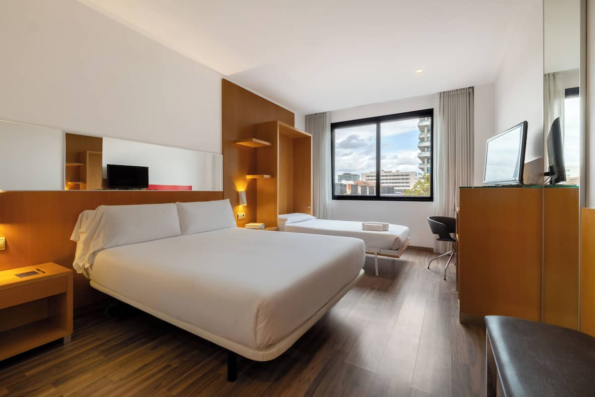Ein Hotelzimmer mit einem großen Bett, einem Einzelbett, einem Schreibtisch, einem Sessel, einem Fernseher und einem Fenster mit Blick auf die Stadt.