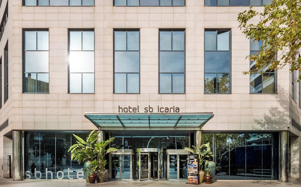 Entrada de hotel con fachada de vidrio, zona de acceso cubierta y plantas en macetas.