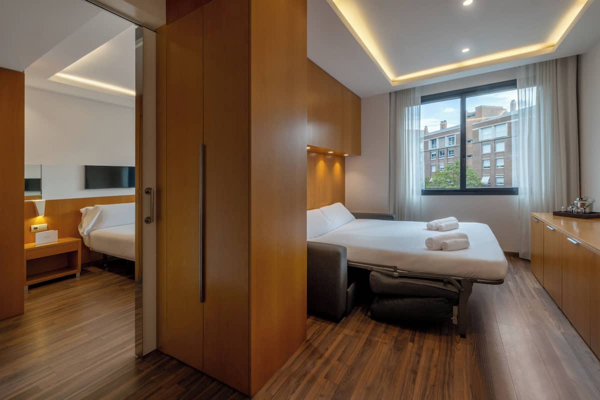 Ein Hotelzimmer mit Bett, Schreibtisch, Stuhl, Kleiderschrank und einem Fenster mit Blick auf Gebäude.