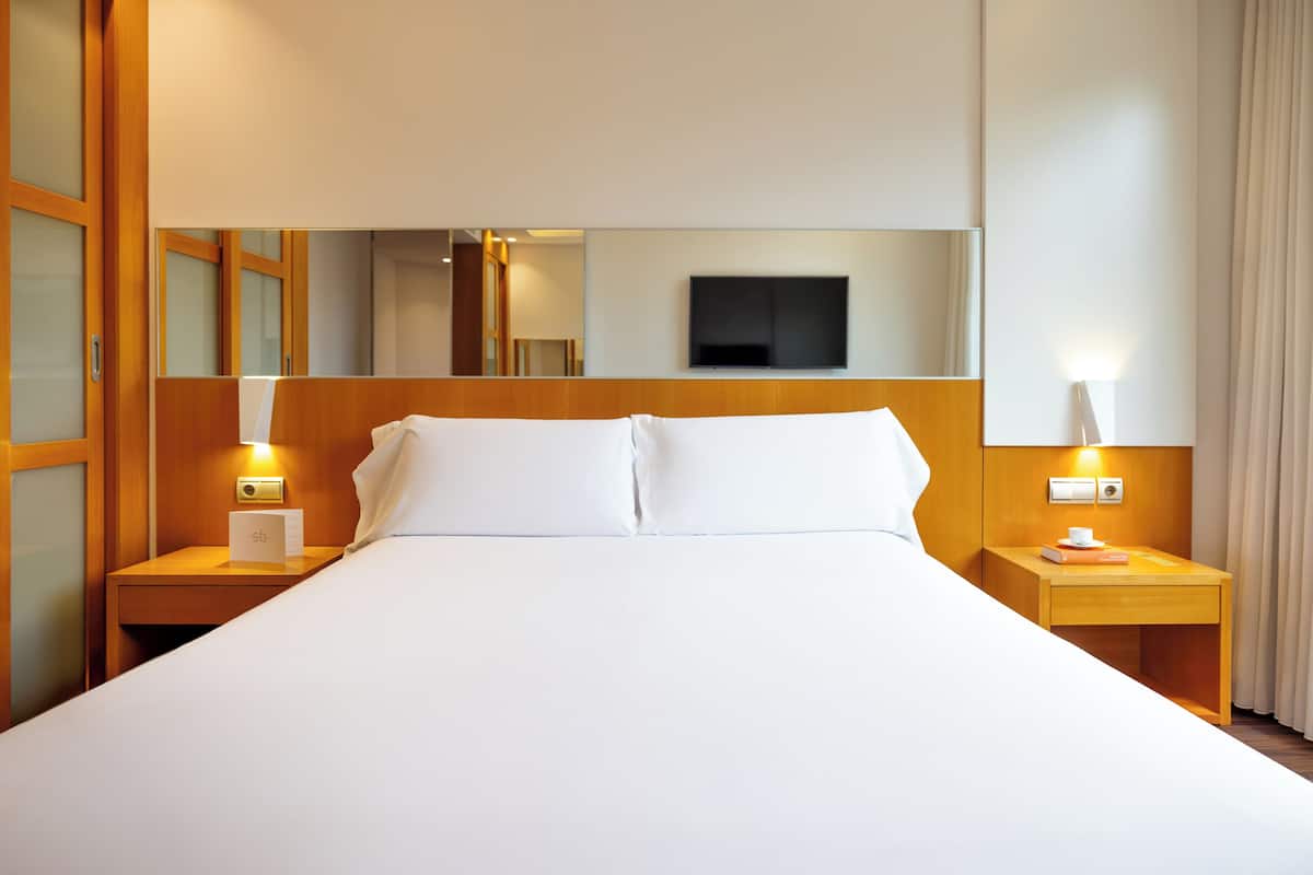 Ein Hotelzimmer mit einem großen Bett, einem Holzkopfteil, Nachttischen, einem Flachbildfernseher und einem Spiegel.