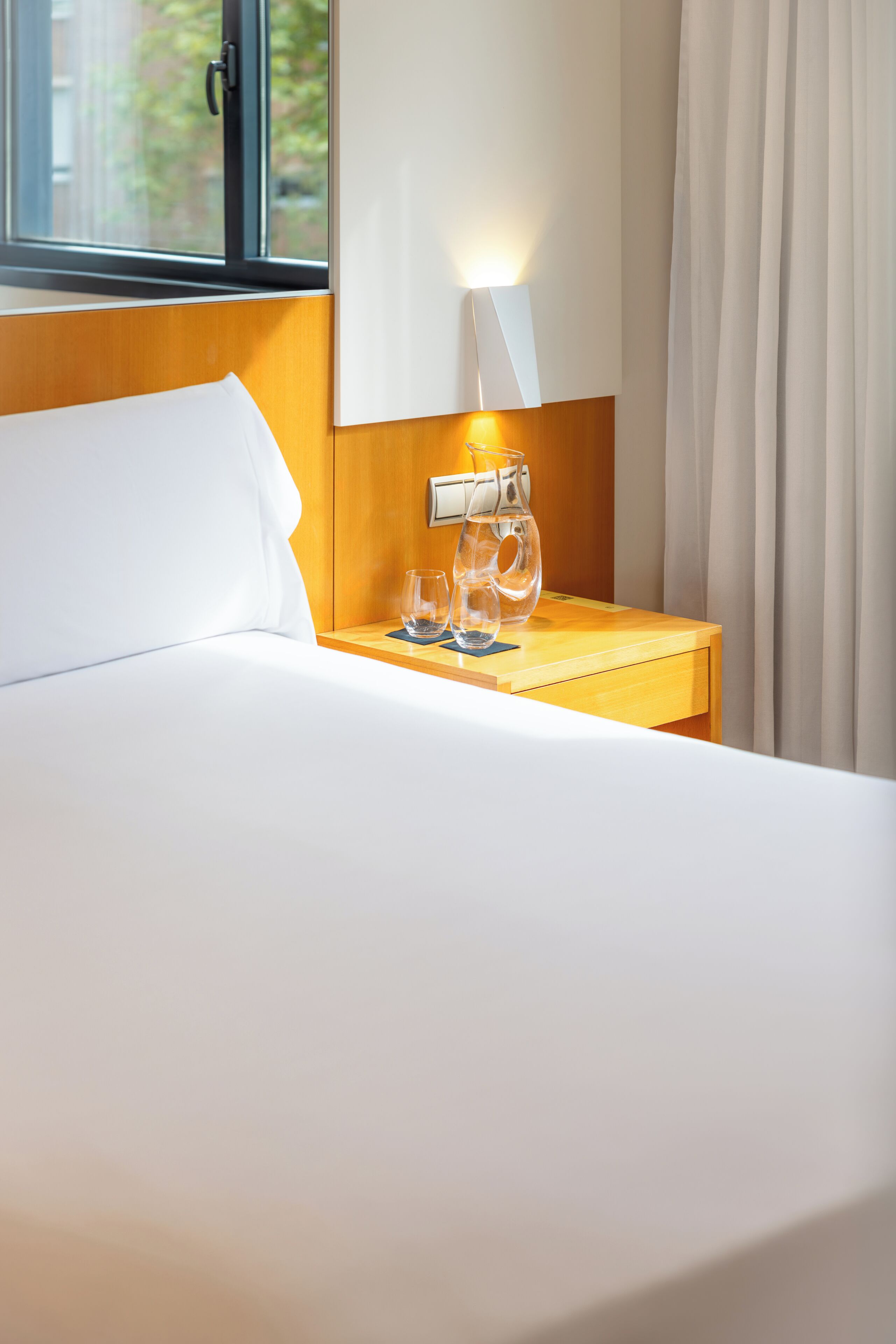 Een hotelkamer met een bed, een nachtkastje met een lamp en een raam met gordijnen.