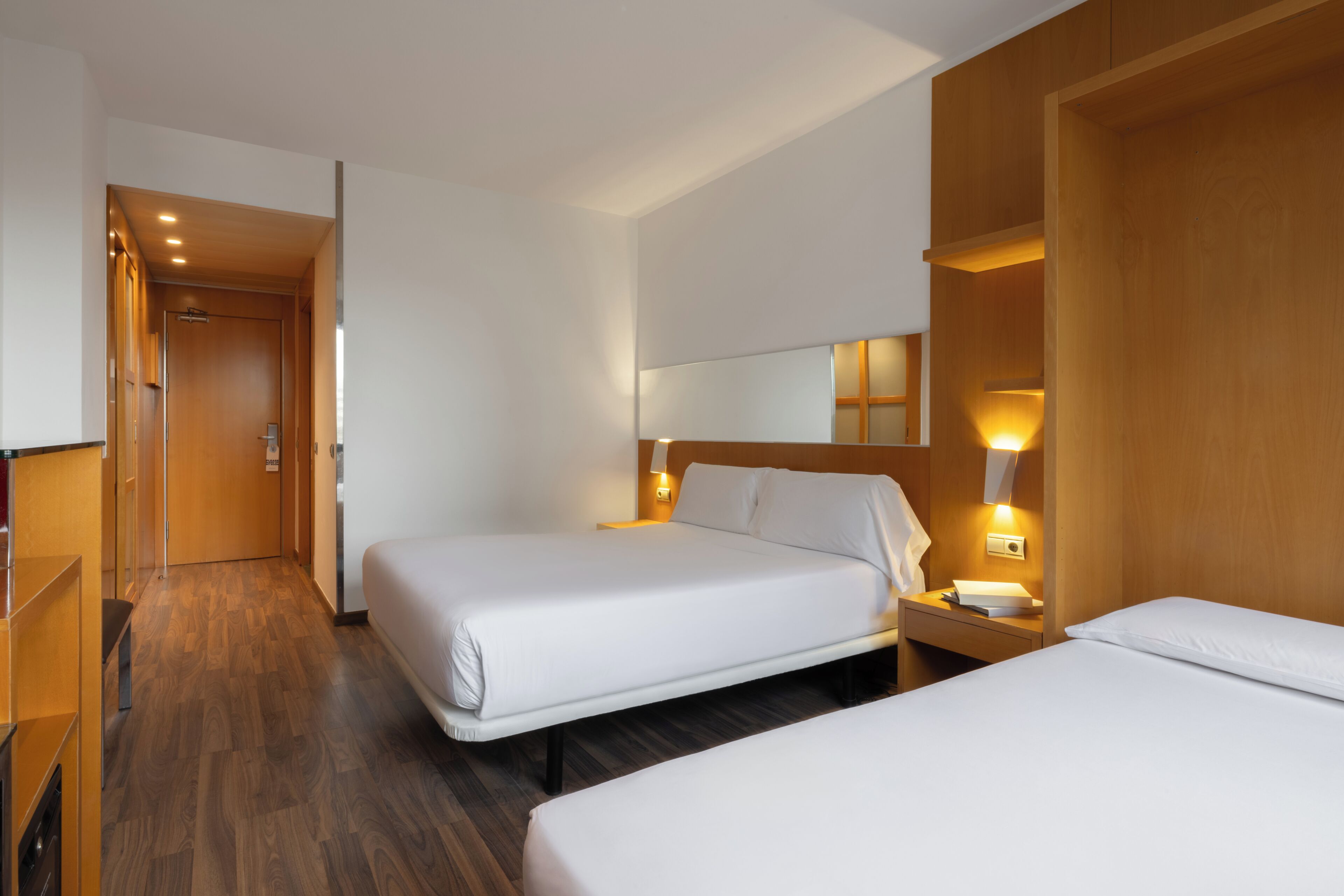 Habitación de hotel con dos camas, suelo de madera, un armario de madera y una mesita de noche con lámpara.